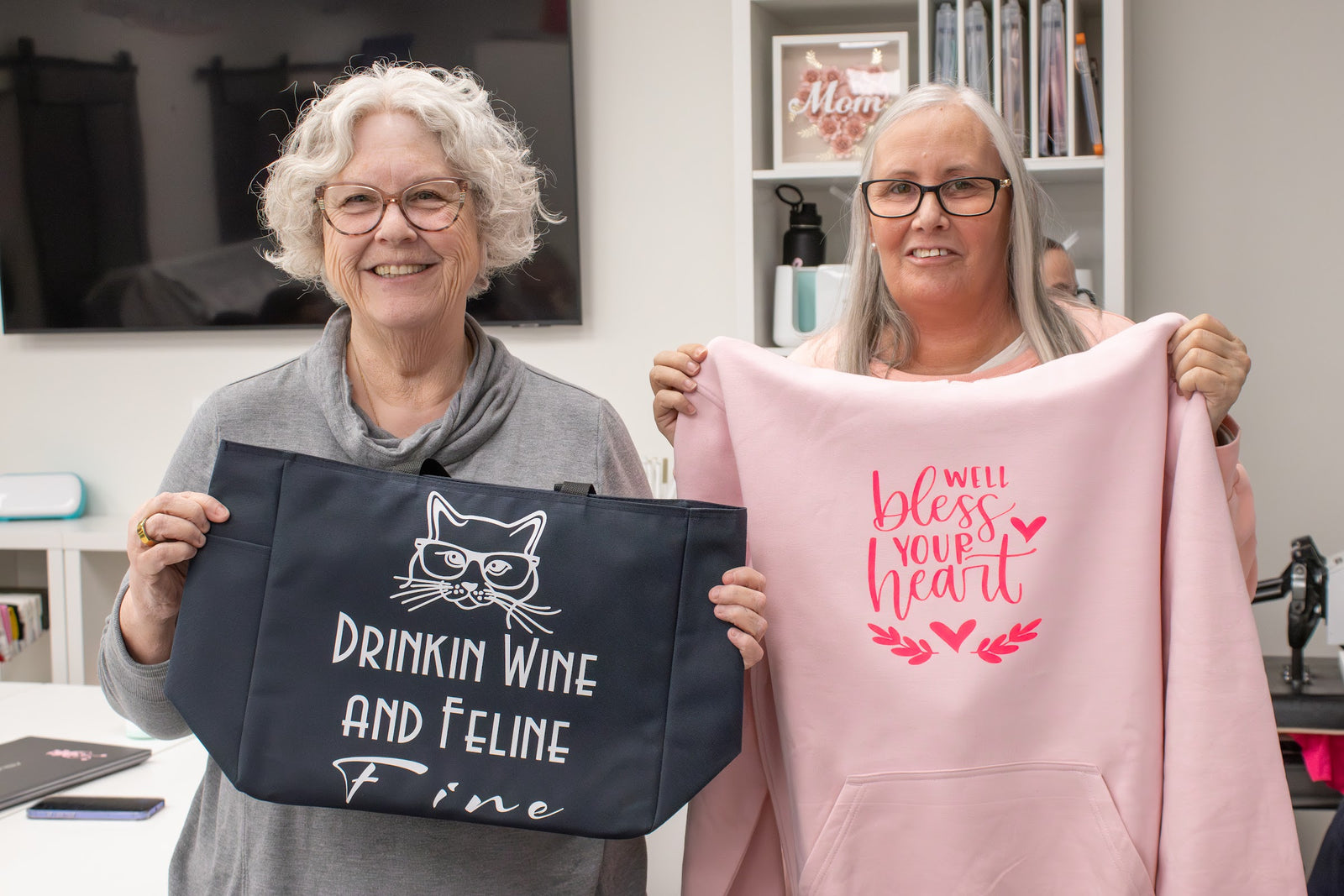 Two smiling makers holding up the tote bag and hoodie they made at a Uniquely Whynot Craft workshop